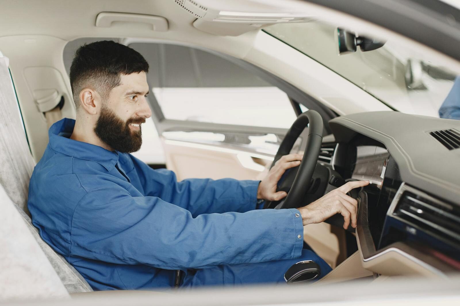 Mechanic in blue coveralls interacts with car dashboard, smiling and focused.